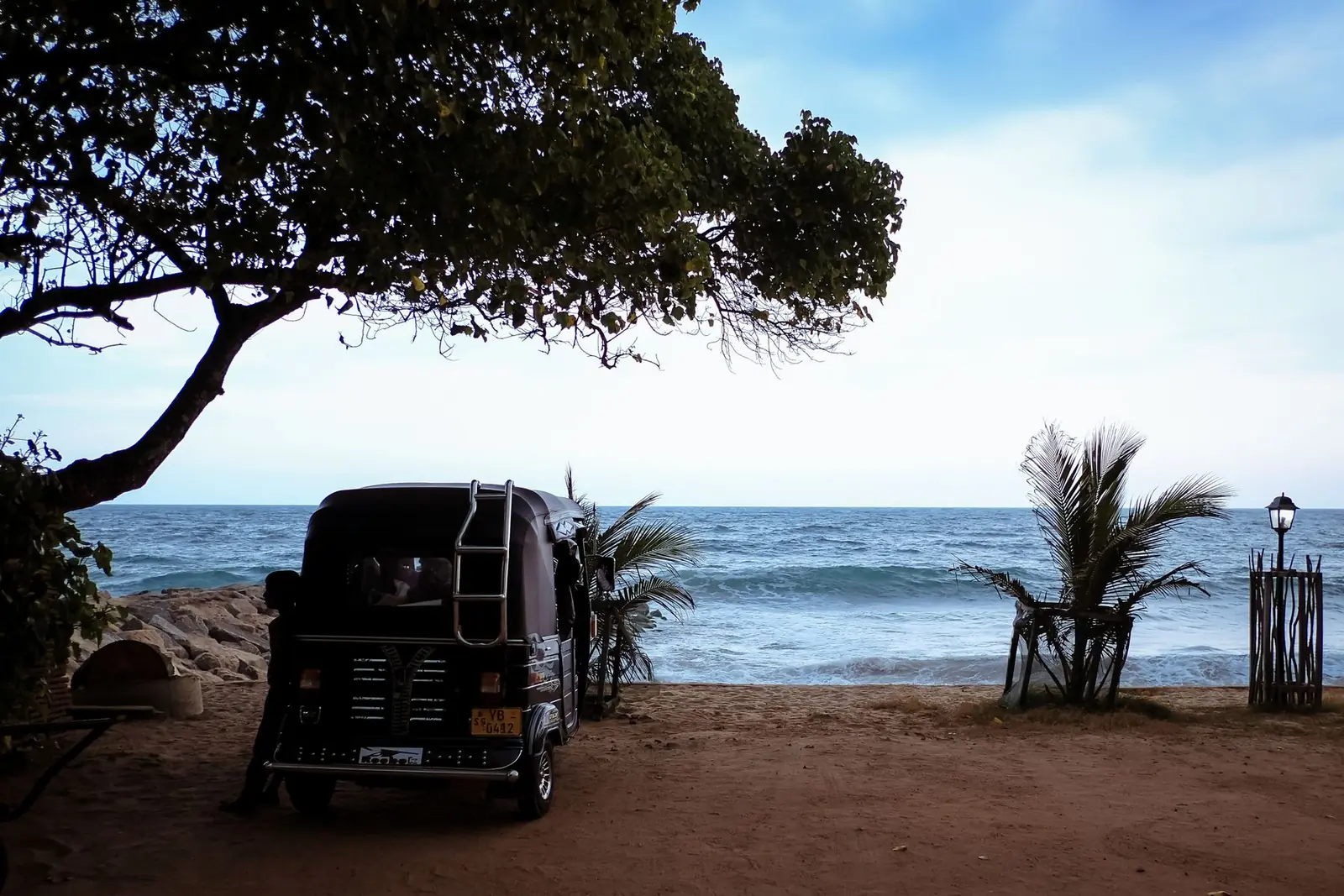 A Sri Lanka tuk tuk parked under a leafy tree beside a west coast beach