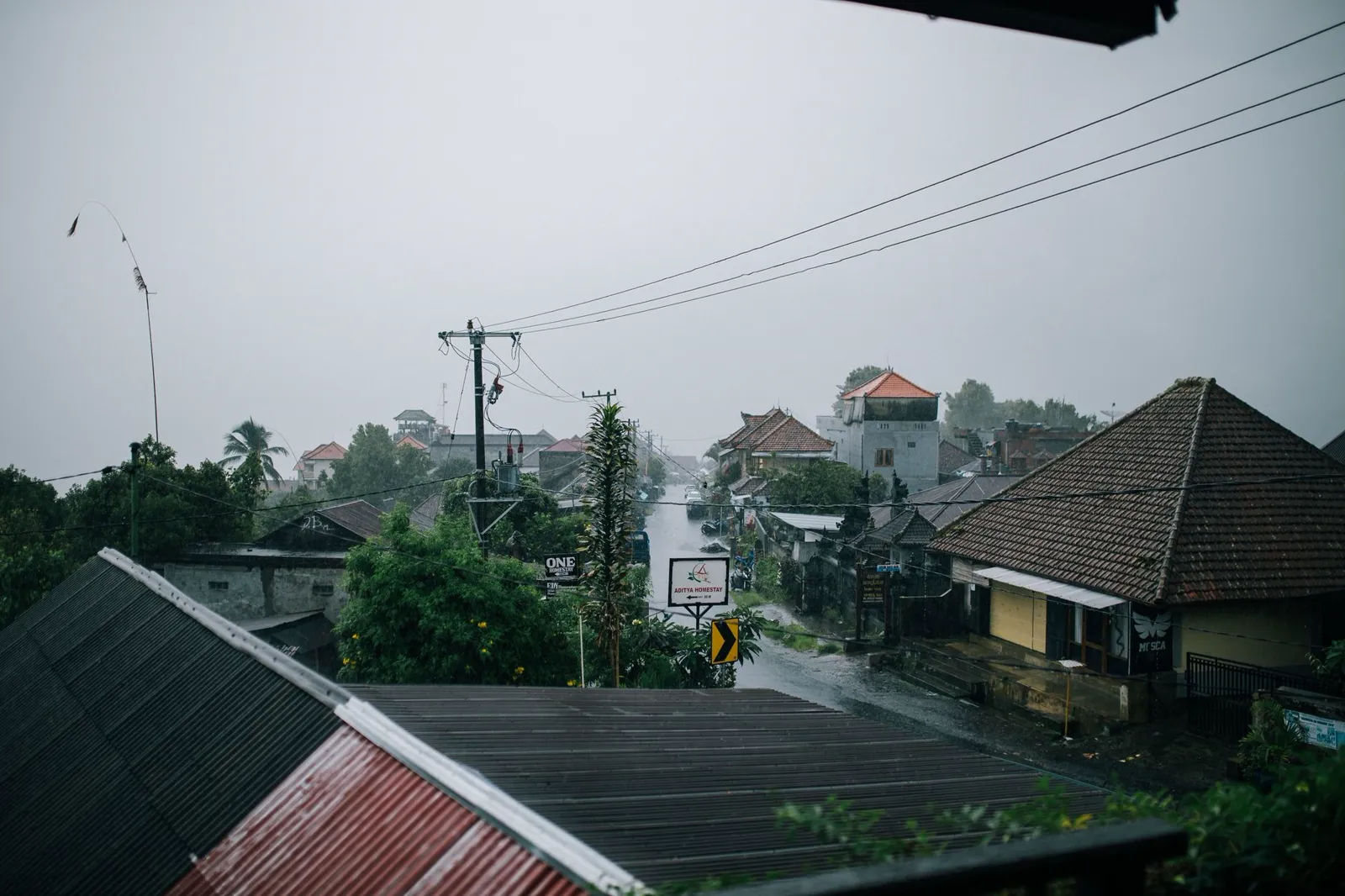 A wet street under a flat grey sky, with rain lying on the road between tiled roofs